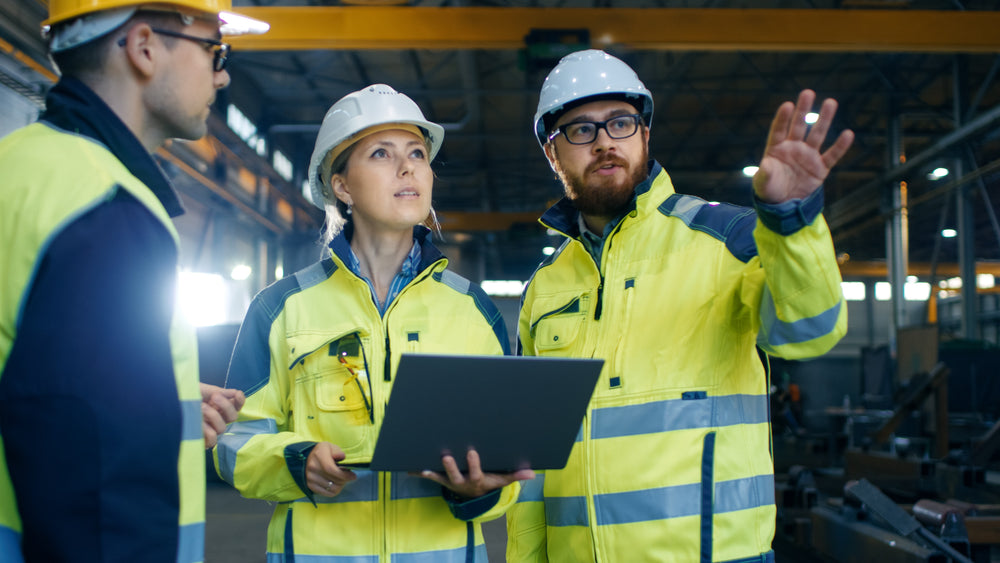 three young workers assess a construction site, female worker holds a laptop as they review plans